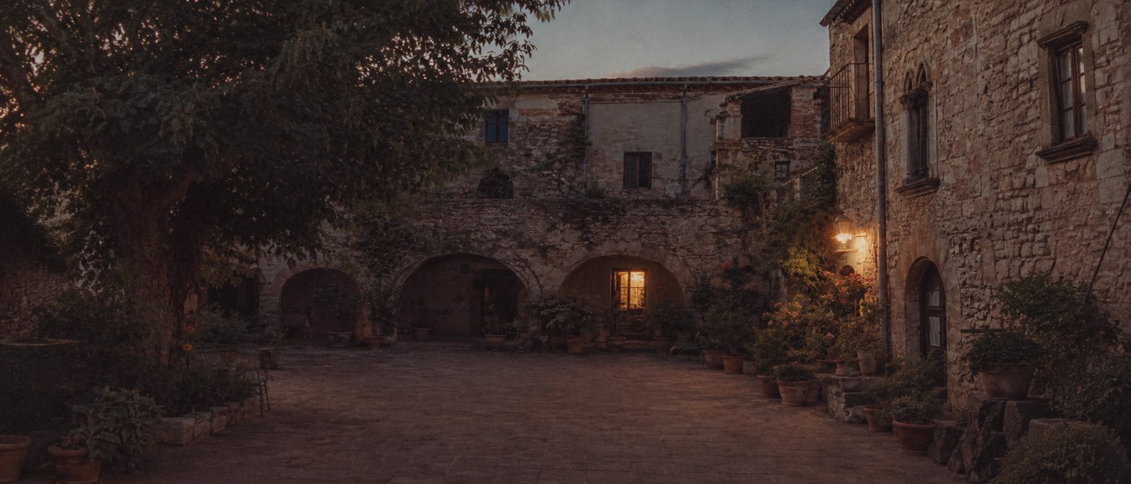 A stone courtyard at dusk, a lit window and a lantern glowing